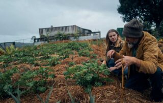 A young woman and an older man crouch down next to a field of crops as the man explains something to the woman