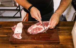 A large piece of red meat is being trimmed by a knife by a man wearing a black watch