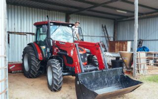 A large red tractor and a man with a baseball cap standing at the door of the tractor, smiling