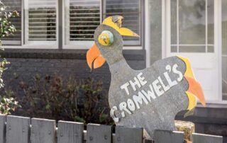 A dark grey fence with a fake chicken sign made out of metal that says The Cromwells in silver lettering