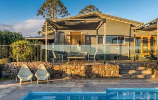 A grey house with a deck that leads out to a blue swimming pool with two sun lounger chairs next to it