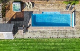 A bird's eye view of a long, blue, rectangular pool, a hot tub and sun lounger chairs alongside a green yard
