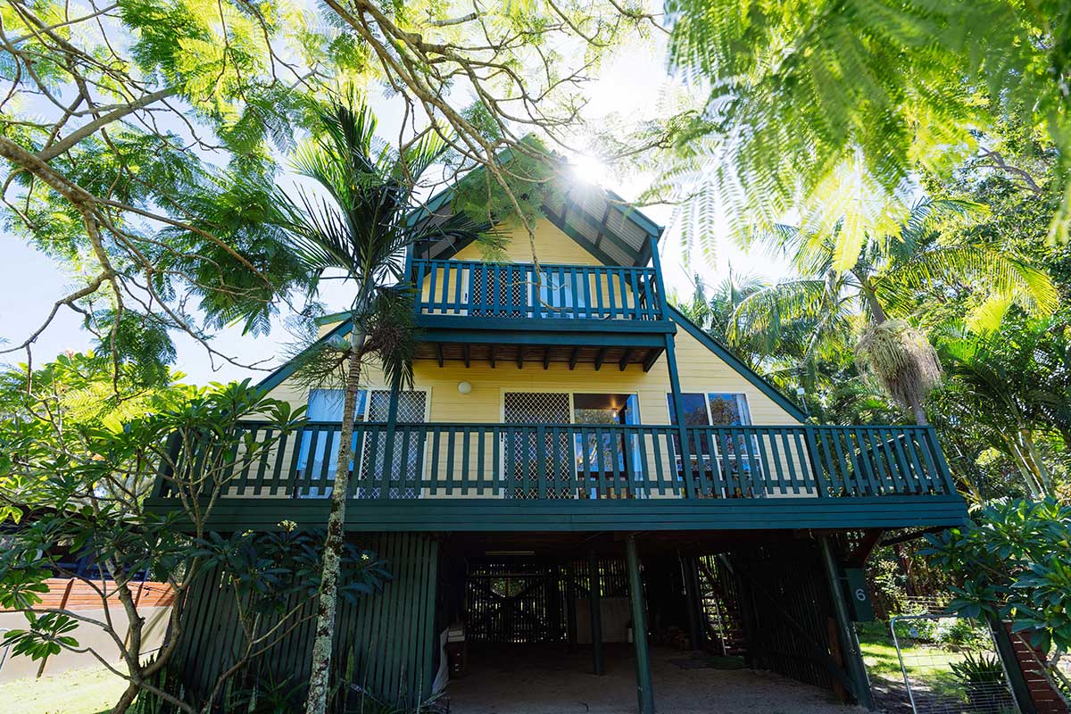 A two-storey, yellow A-frame house with a dark green deck and palm trees