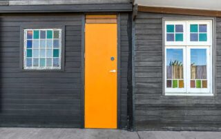 Middle Shed front door that is bright orange and have two windows of coloured glass on either side. The house is dark brown/black
