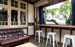 Middle Shed lounge room with a dark leather couch and three white stools at a breakfast bar with a view outside to a green tree