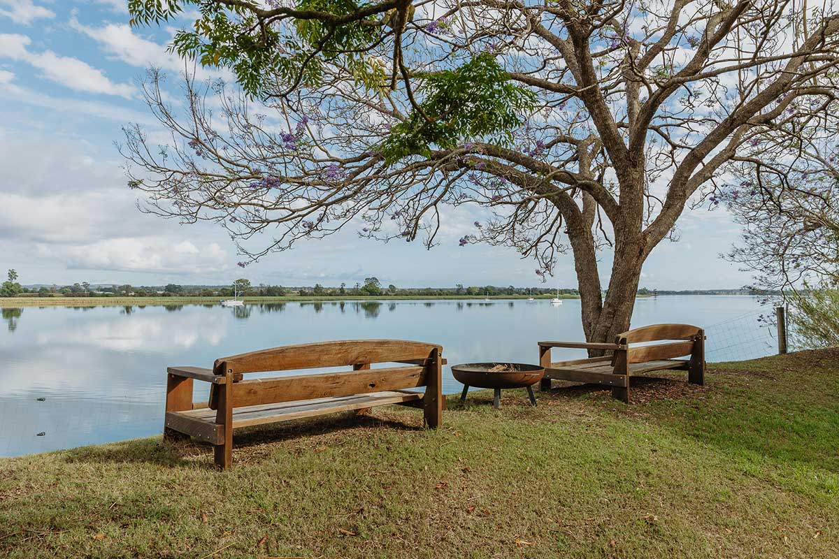 Two wooden benches with a fire pit between them, next to a tree overlooking the Clarence River at Big River Retreat, Ulmarra