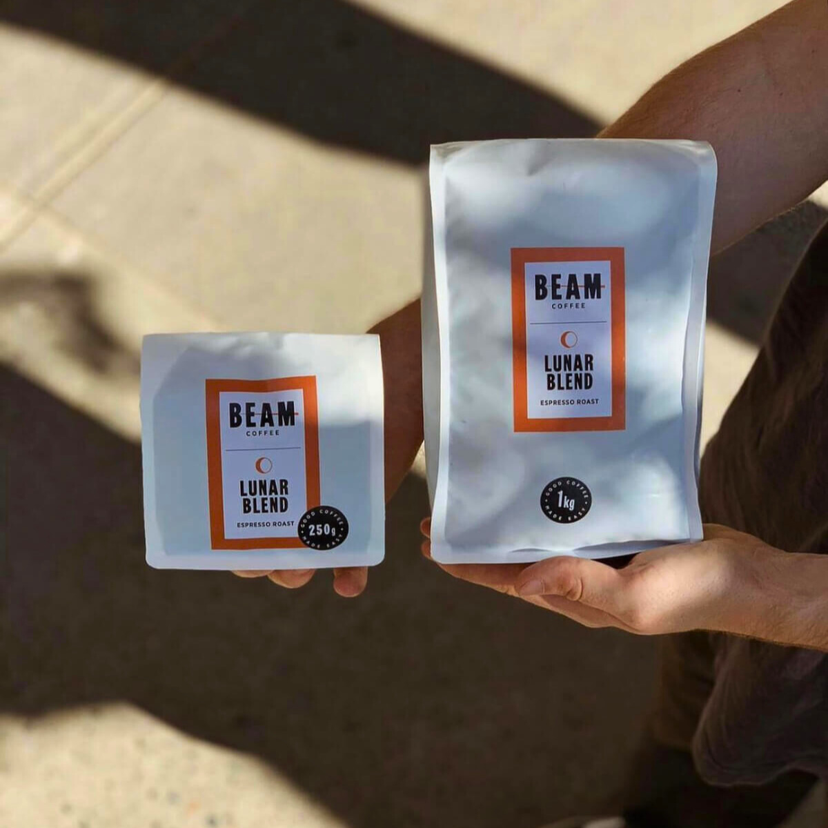 Two hands hold two bags of Beam Coffee beans, one smaller bag and one is larger. The bags are white with Luna Blend on them and an orange border