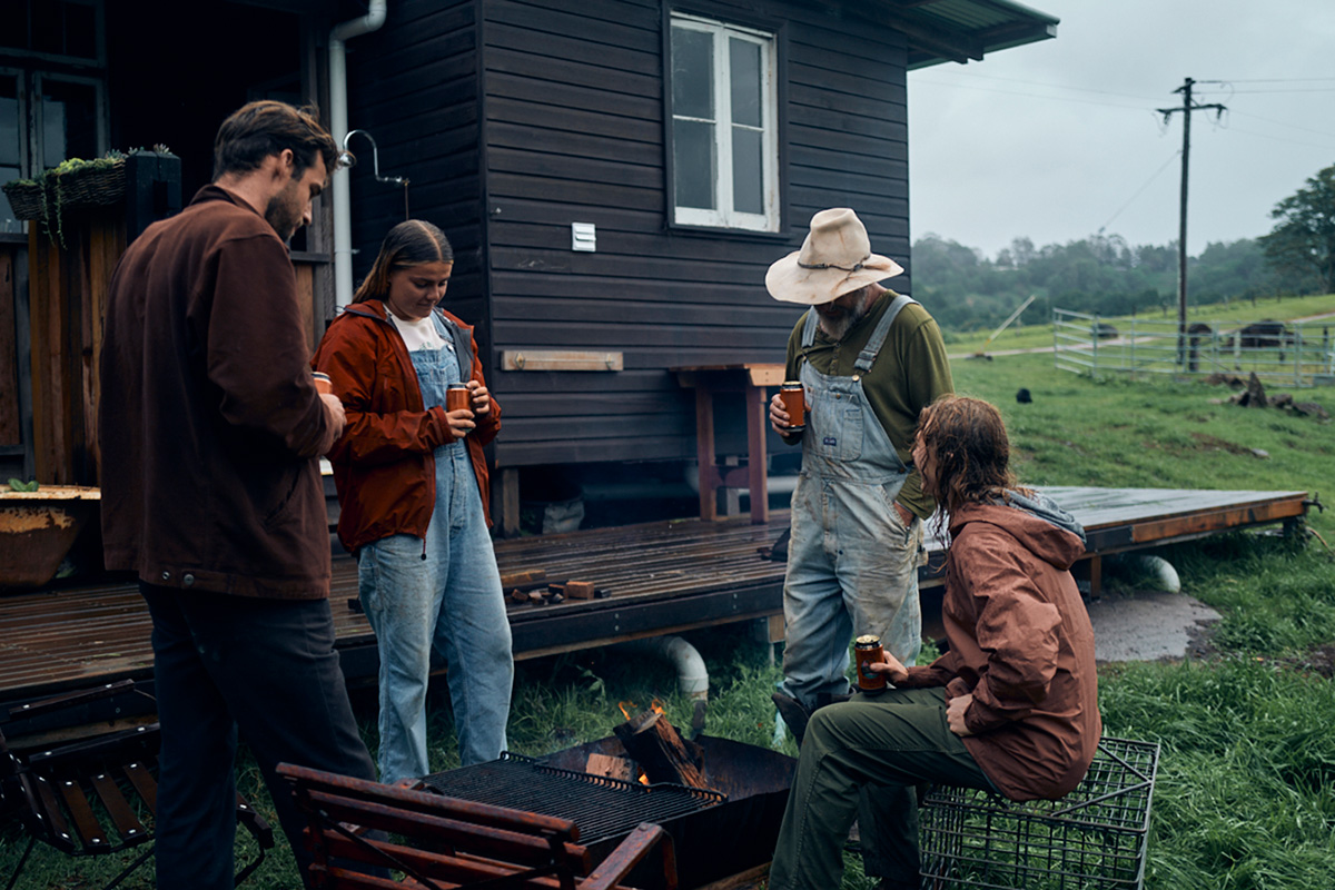 Four people stand around a firepit holding tins of beer on a rainy day. There is a black house behind them with a wooden deck