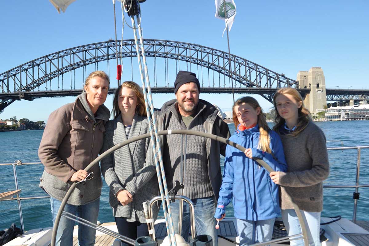 A family of three teenage girls and a man and woman stand on a boat on a sunny day with the Sydney Harbour Bridge in the background