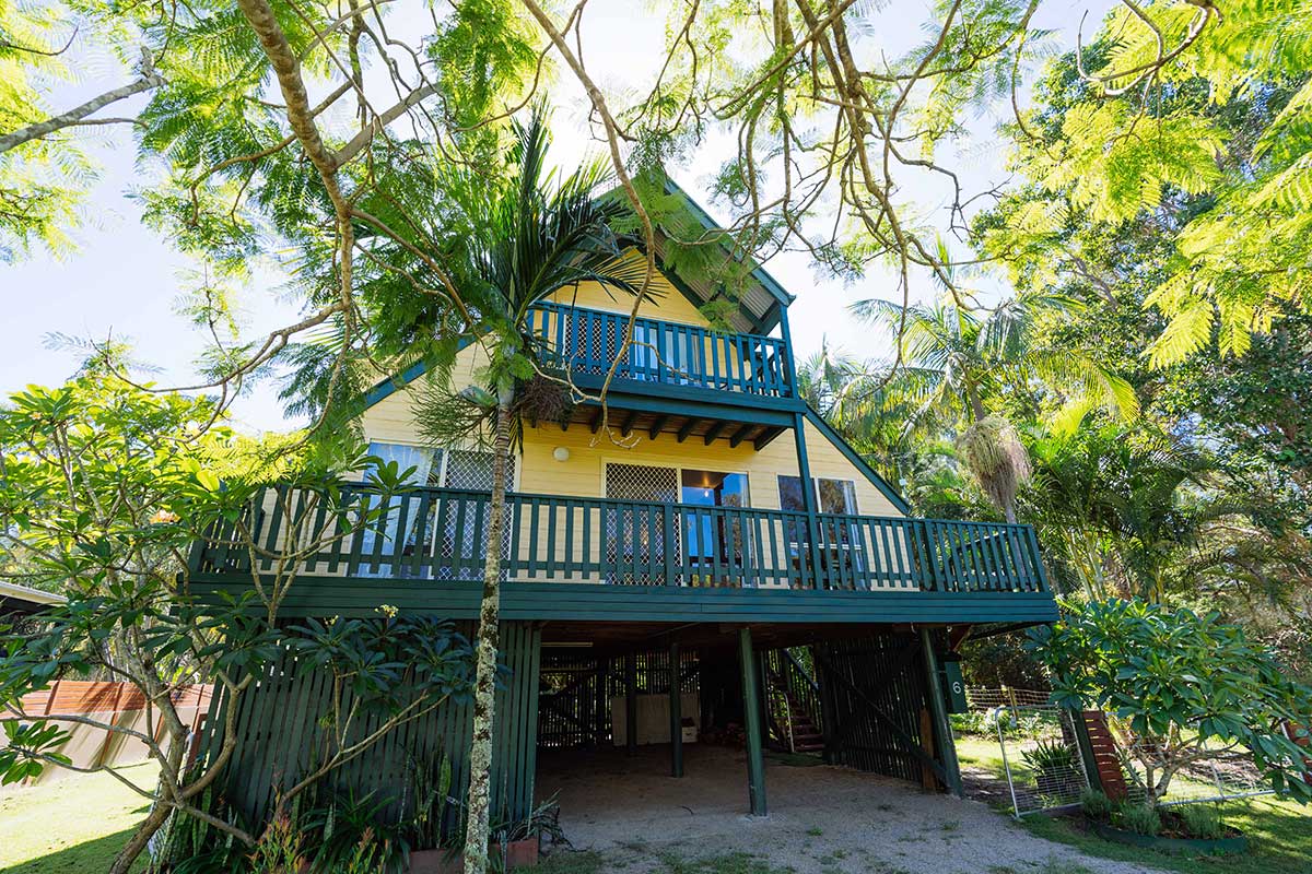 A two-storey, yellow A-frame house with a dark green deck and palm trees