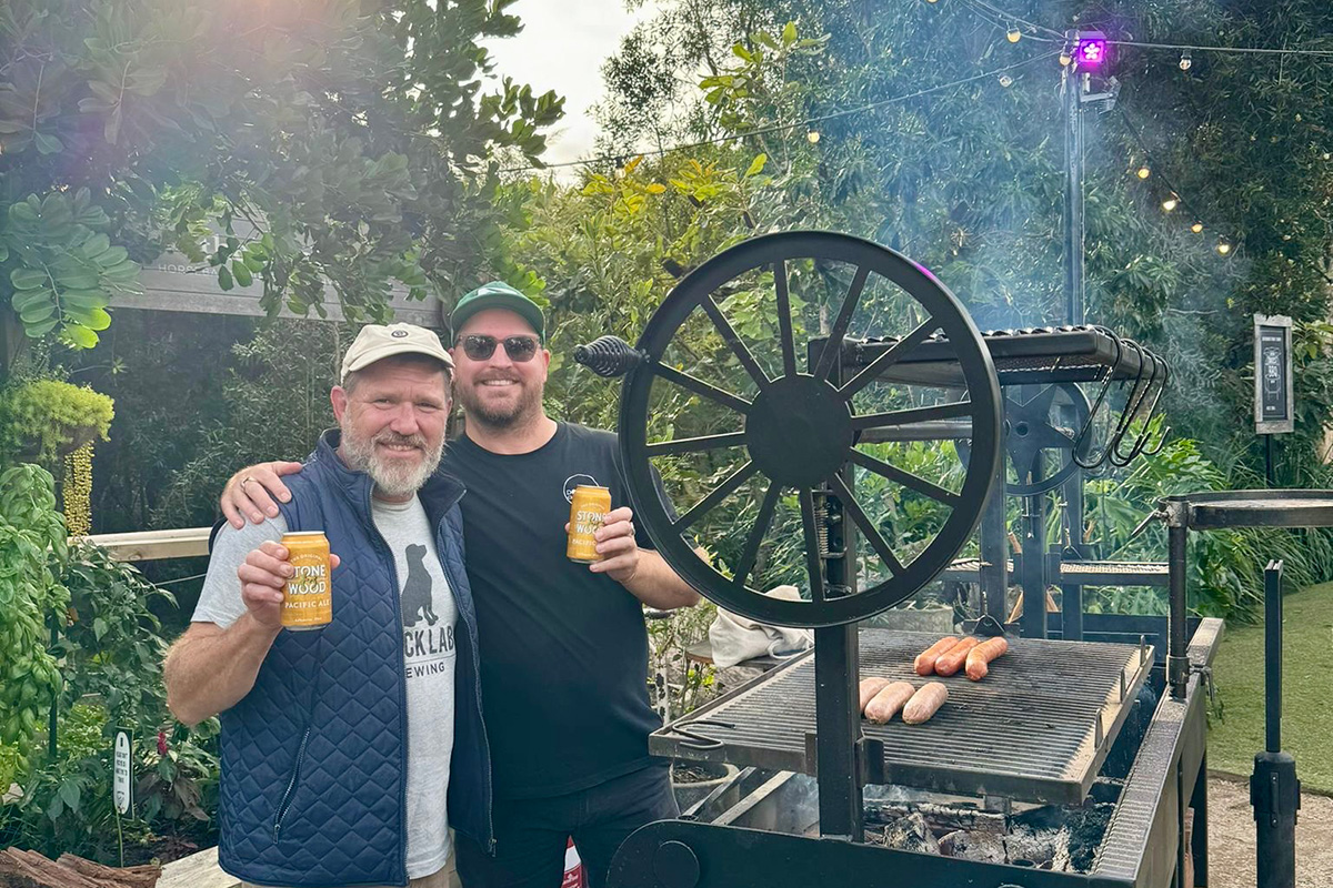 Greg Cromwell and David Lovett standing next to a black bbq grill smiling at the camera and holding up Stone and Woods Pacific Ale tins